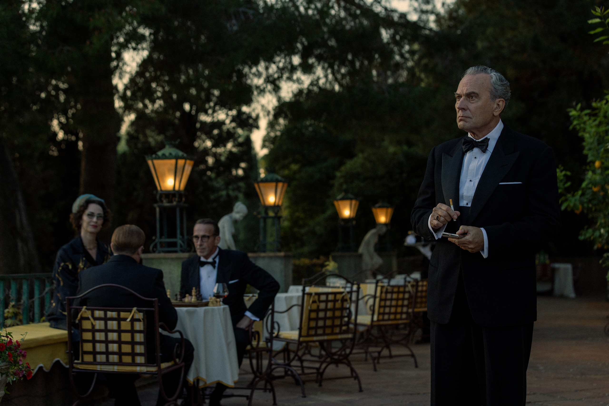 Official Netflix still from The Final Problem: Jose Coronado as Basil on a lantern-lit terrace while guests play chess. Netflix.