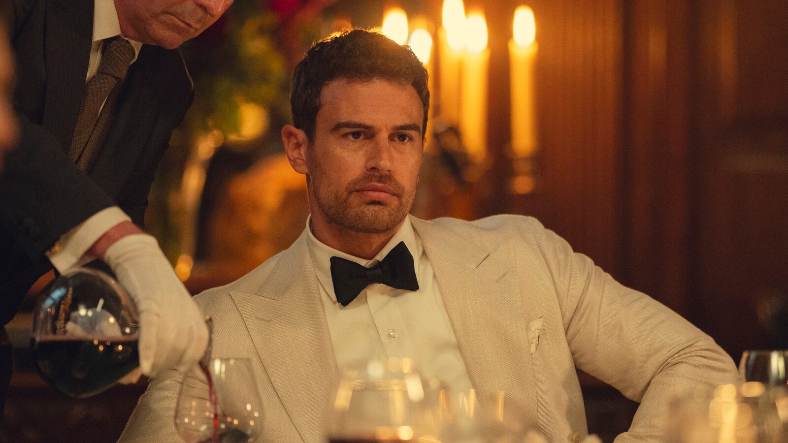 Official Netflix still from The Gentlemen Season 2. Theo James as Eddie Horniman sits in a cream dinner jacket while a white-gloved server pours wine. Courtesy Netflix.