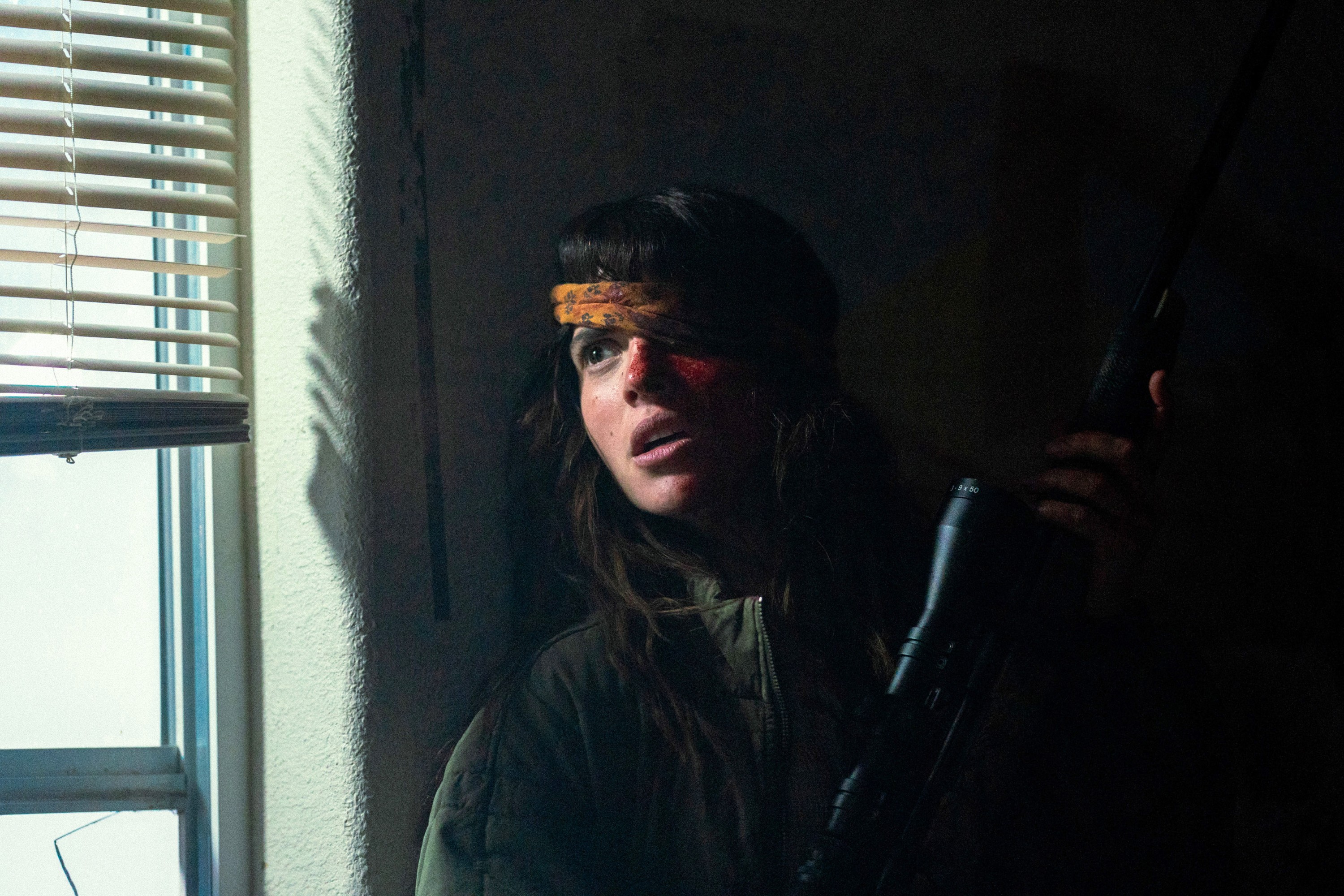 Official still from A24's Onslaught. Adria Arjona as Celeste with the rifle by the blinds. Photo via IndieWire / A24.