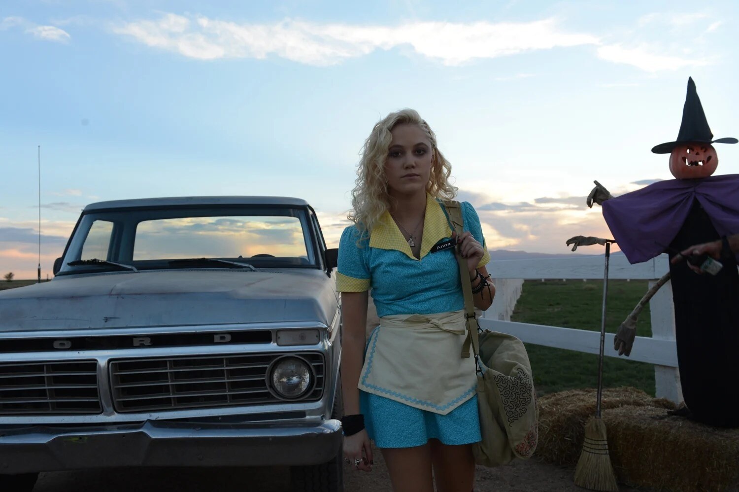Official still from Adam Wingard's The Guest (2014): Maika Monroe as Anna Peterson in waitress blues beside Halloween scarecrow and pickup. The Guest / Picturehouse.
